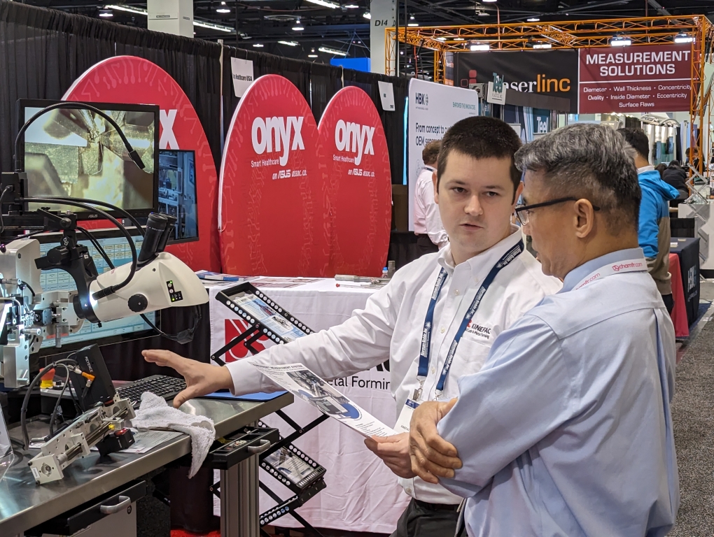 A KineCoil sales engineer in a white shirt demonstrates the MicroCoiler's high-precision coiling capabilities to an expo attendee.