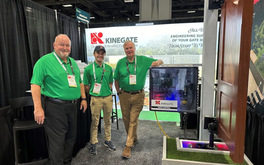 Three men in green KineGate polo shirts stand ready in the KineGate booth. Next to them is a freestanding wooden security gate used to demonstrate KineGate property security solutions.