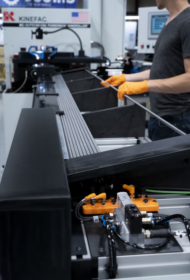 A worker wearing orange gloves loads material into the Kinemat Bar Load System for automated bar feed.